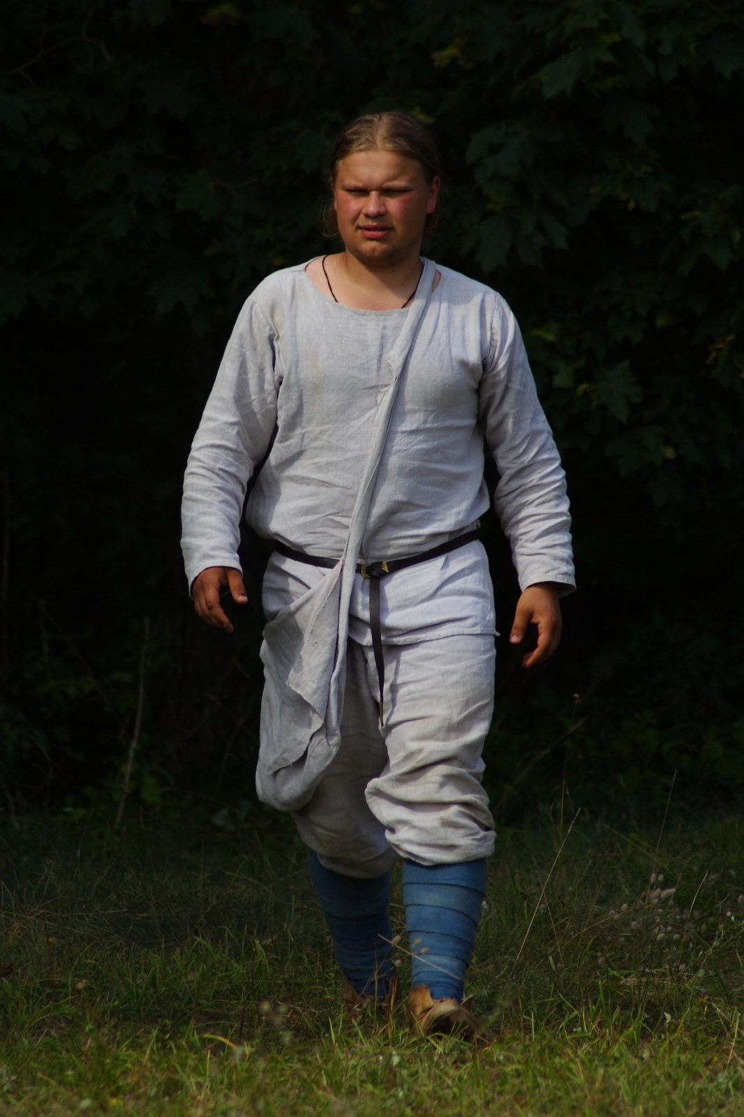 A person, dressed in medieval-style clothing, is walking outdoors during the 2009 Semigallian Days. They wear a loose, long-sleeved white tunic with a belt, white breeches, and blue leg wraps. The background is dark green foliage. The lighting suggests a natural setting with a mix of sunlight and shadow.