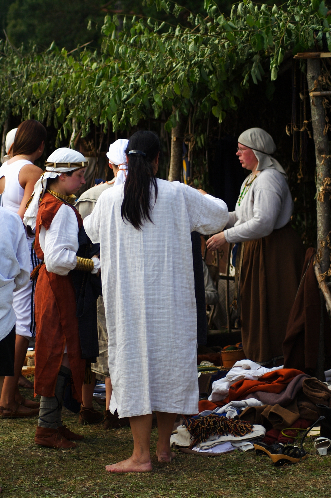 A group of people, some in historical or medieval attire, are at an outdoor event under a leafy canopy. One person in a long white robe and bare feet stands with their back to the camera. Others wear various period costumes, including head coverings and tunics, reminiscent of the 2009 Semigallian festival.