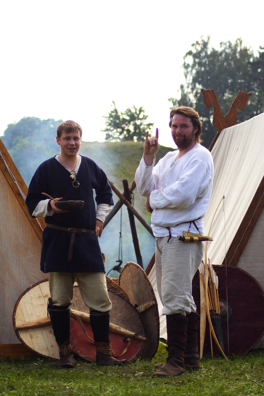 Two men dressed in medieval-style clothing stand in front of canvas tents at the Semigallian Days cultural event. The man on the left, in a navy tunic and beige trousers, holds a wooden bowl. The man on the right, in a white tunic and trousers, holds up a small purple object. Shields rest against the tents behind them.