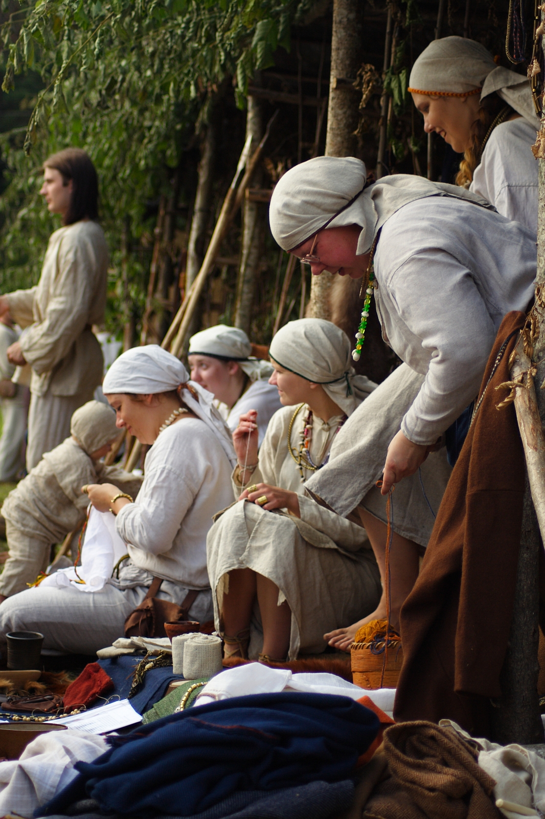 A group of individuals dressed in traditional, medieval-style clothing sits and stands near a wooden structure with greenery at the Semigallian festival. Some are engaged in crafts such as embroidery and braiding. Most are wearing light-colored garments and head coverings, creating a historical and communal atmosphere.