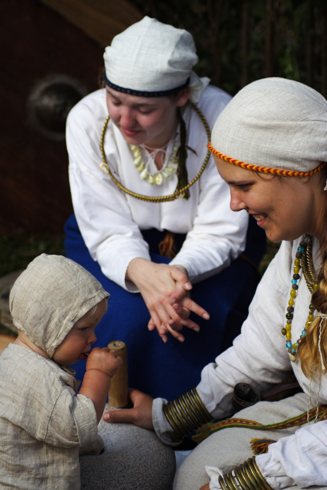 Two women in traditional clothing and headscarves sit on the ground with a baby, who is also dressed in traditional attire. The woman on the right smiles at the baby, while the other woman looks on. Their close interaction suggests they are part of a family or community gathering at a Semigallian festival.