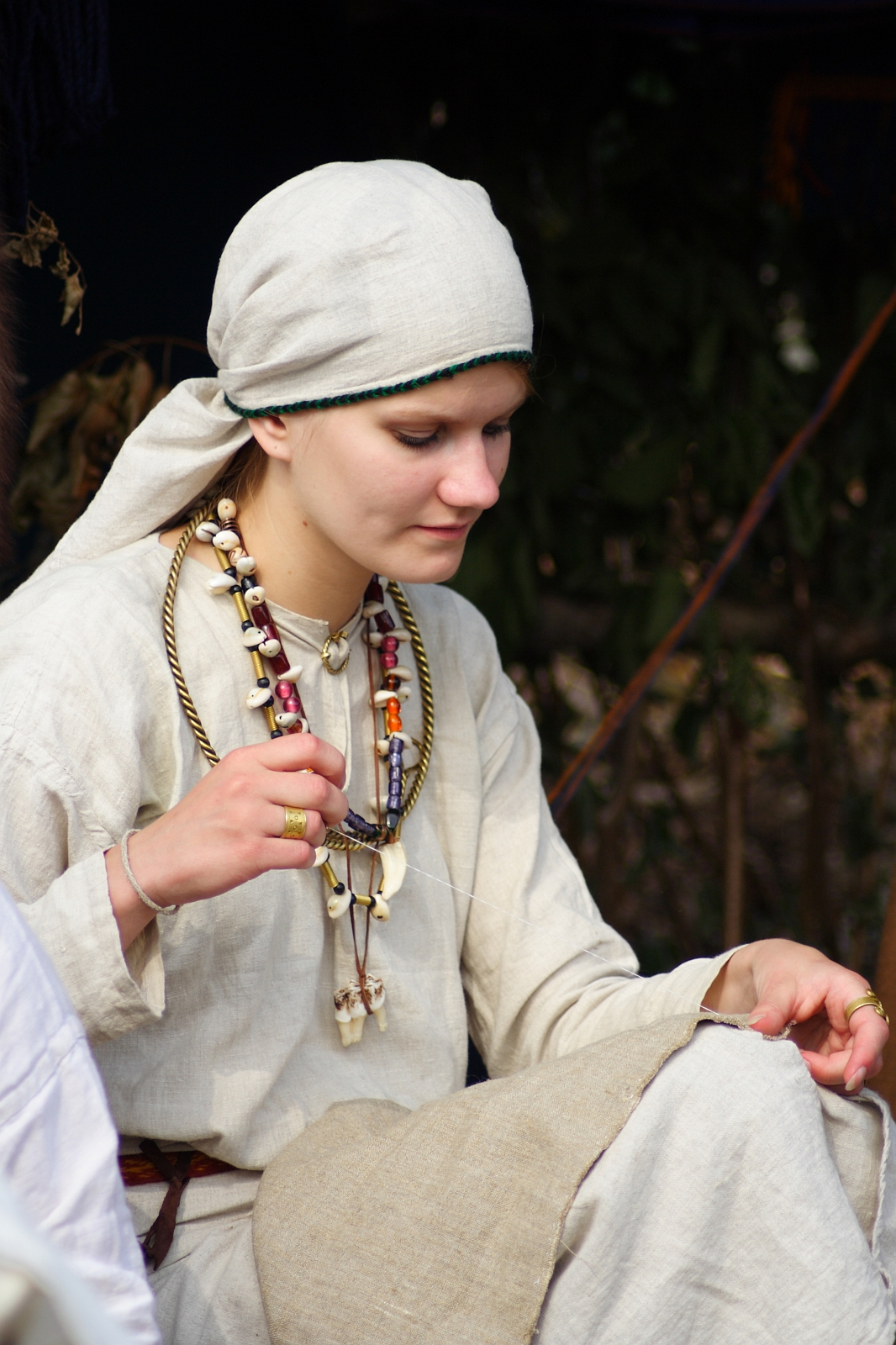 A person dressed in historical attire, wearing a beige headscarf and matching tunic, sits with a focused expression at the Semigallian Days festival. They have multiple necklaces with various beads and ornaments. Their right hand rests on their lap, holding part of their garment, while the background is blurred with greenery.