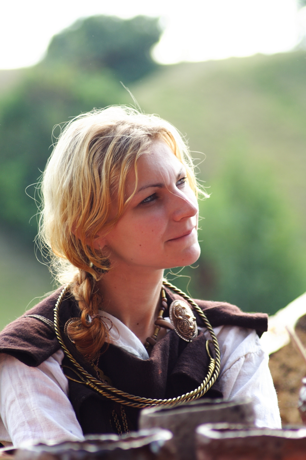 A woman with blonde hair braided on one side is dressed in historical attire, including a dark shawl, a large ornate brooch, and a twisted necklace. She is outdoors at the Semigallian festival, with blurred greenery in the background, and gazes thoughtfully to her right, bathed in soft natural light.