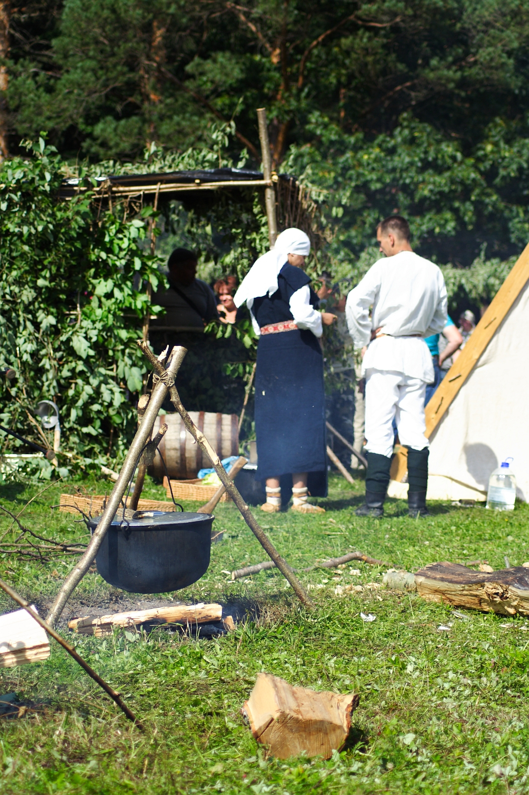 A historical reenactment scene set outdoors. In the foreground, a black cauldron hangs over a fire tripod with logs nearby. Two people in period clothing, one in a dark dress with a white headscarf and the other in a white tunic and black boots, stand conversing near a green bush during Semigallian Days 2009.