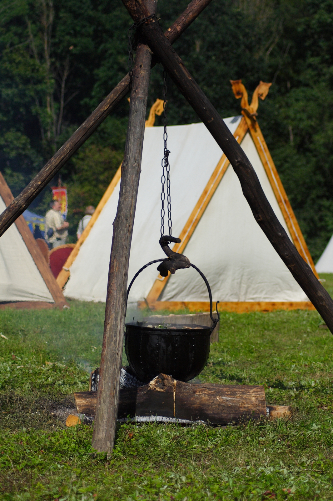 A tripod made of sturdy wooden poles supports a hanging cauldron over a small fire. The cauldron is suspended by a chain and sits above a partially burned log. In the background, white canvas tents with wooden frames are visible amidst green foliage, evoking the rustic charm of the 2009 Semigallian festival.