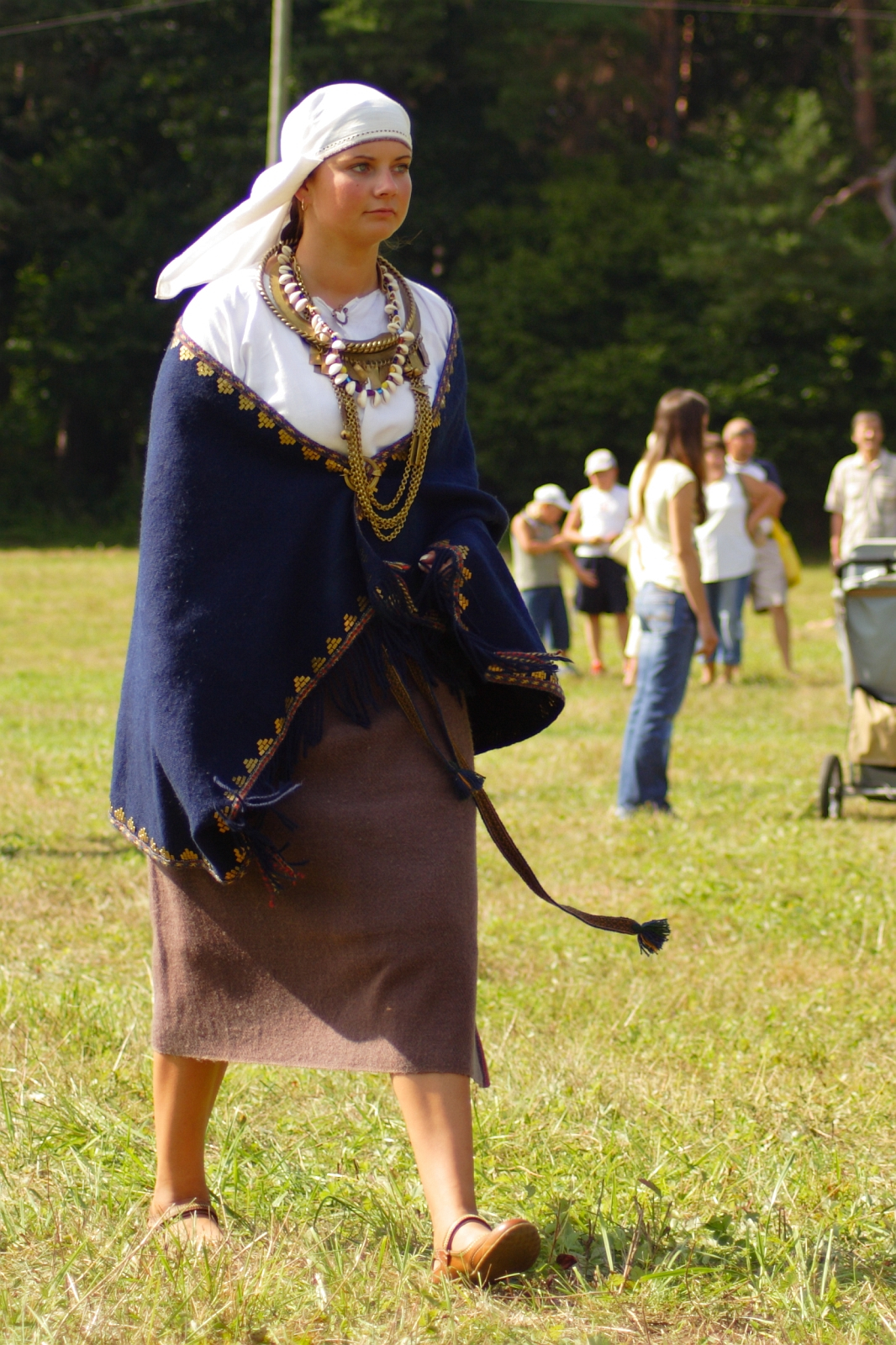 A woman walks on grass, wearing a traditional outfit: a white headscarf, a shawl with gold accents draped over her shoulders, a white top, a brown skirt, and tan shoes. She wears multiple necklaces and holds a strap. The scene is set during Semigallian Days in 2009. People and trees are visible in the background.