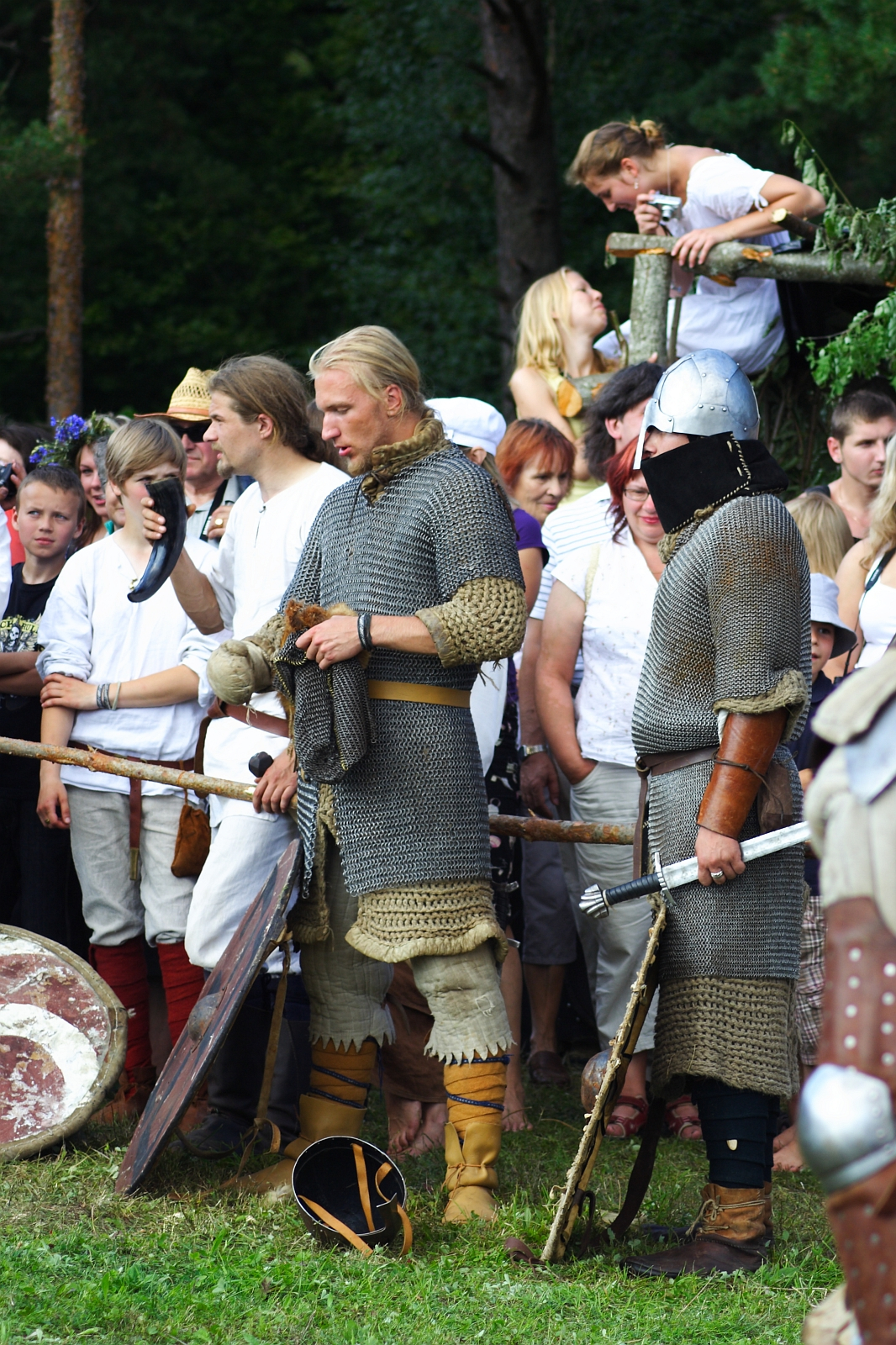 A group of people, some in medieval attire, participate in an outdoor event. In the center, two men dressed as Semigallian warriors in chainmail and leather armor stand in conversation. One holds a sword, and a crowd of onlookers, including children, is gathered behind them. Trees fill the background.