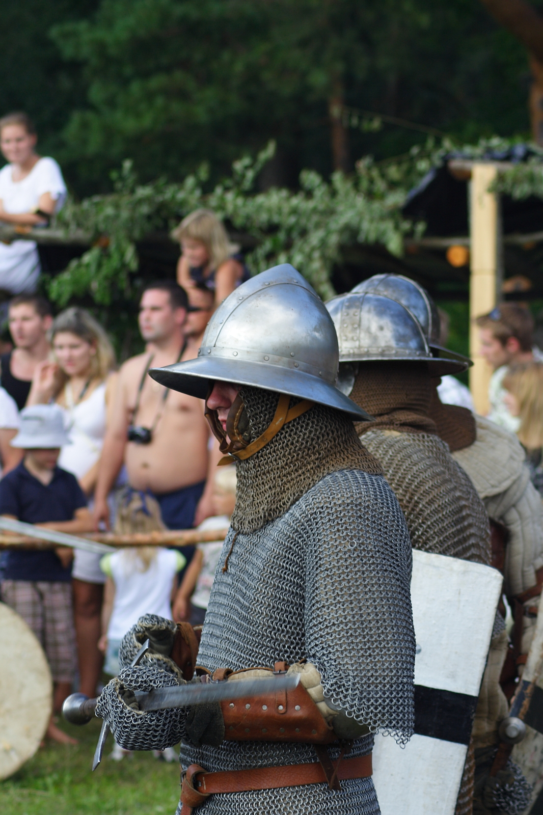 Two medieval reenactors dressed in chain mail and metal helmets stand side by side, holding swords and shields at the Semigallian Days event. They are part of a historical reenactment, watched by a crowd of onlookers in modern clothing. Greenery and wooden structures decorate the background.