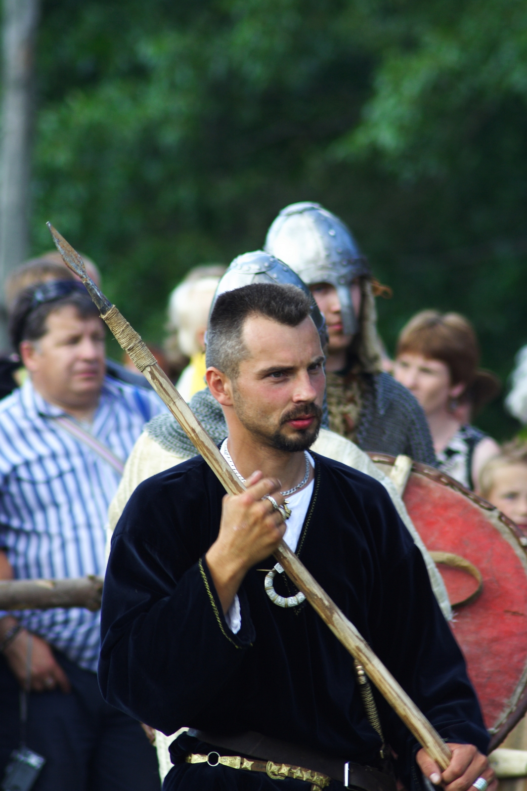 A man dressed in medieval clothing holds a spear, standing among a crowd at Semigallian Days 2009. He wears a dark tunic and has short hair and a beard. Behind him, a man in chainmail armor and a helmet is visible, along with other onlookers, some of whom are dressed in modern clothing. Trees are in the background.