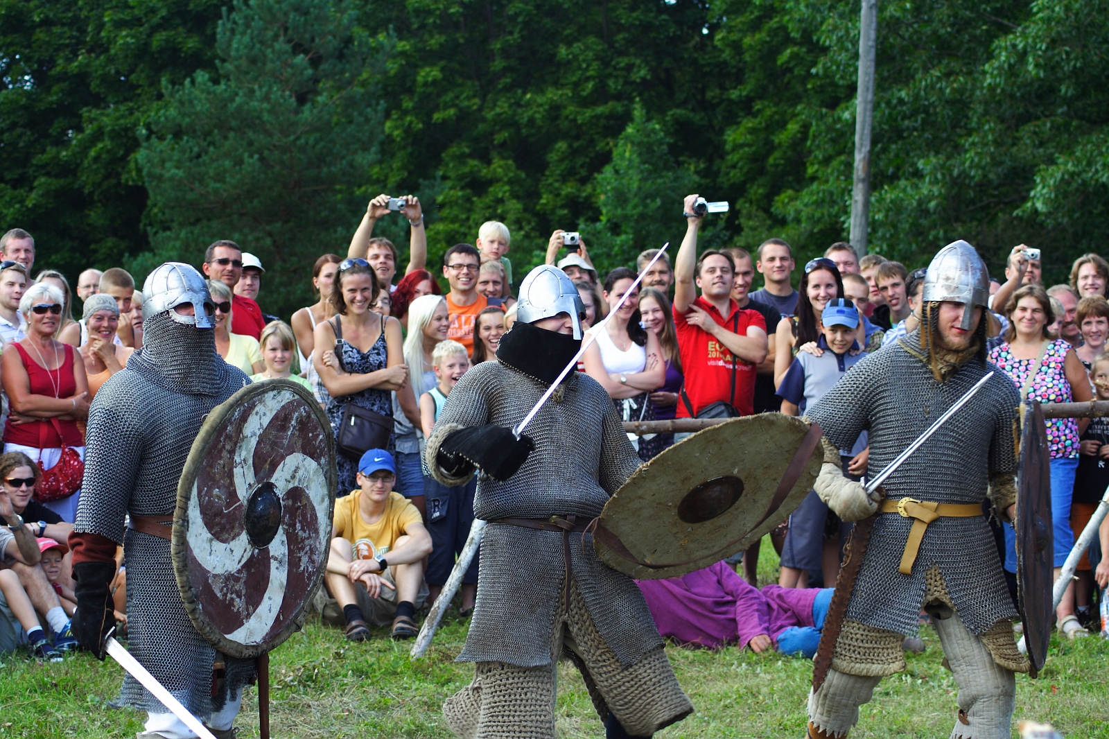 Three people dressed in medieval chainmail armor, helmets, and holding swords and shields, engage in a reenactment during Semigallian Days 09. A large, cheering crowd of men, women, and children look on and take photos. Trees and greenery form the backdrop of this outdoor event.