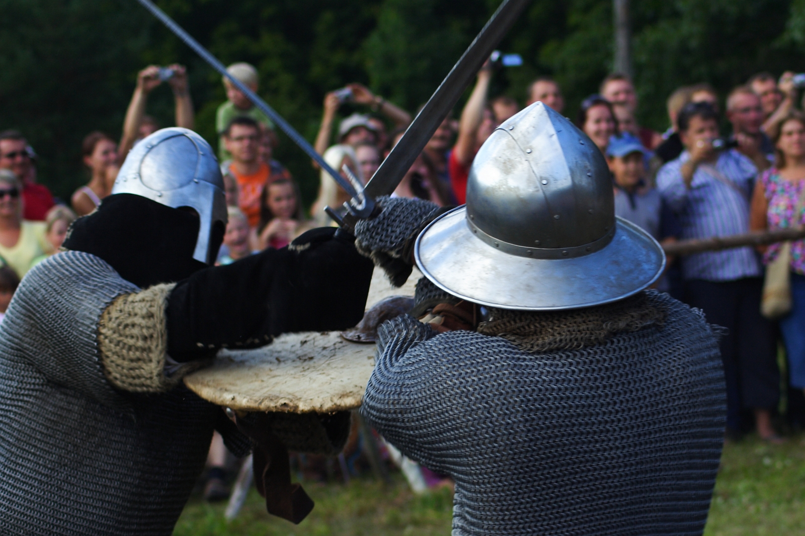 During the Semigallian Days '09, two knights in chainmail and metal helmets engage in sword combat during a reenactment. Both have raised swords clashing together and one carries a large shield. Onlookers in the background, some taking photos, watch the scene set outdoors among trees and greenery.