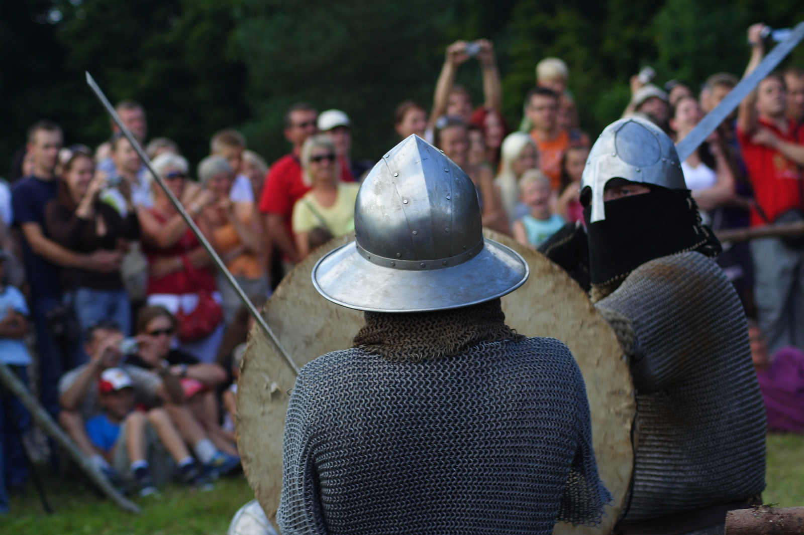 During the 2009 Semigallian Days, two people dressed in medieval armor and chainmail engage in a mock battle, holding swords and shields. They are surrounded by a crowd of spectators in casual attire, eagerly watching the performance, some taking photos. The event appears to be outdoors with trees in the background.