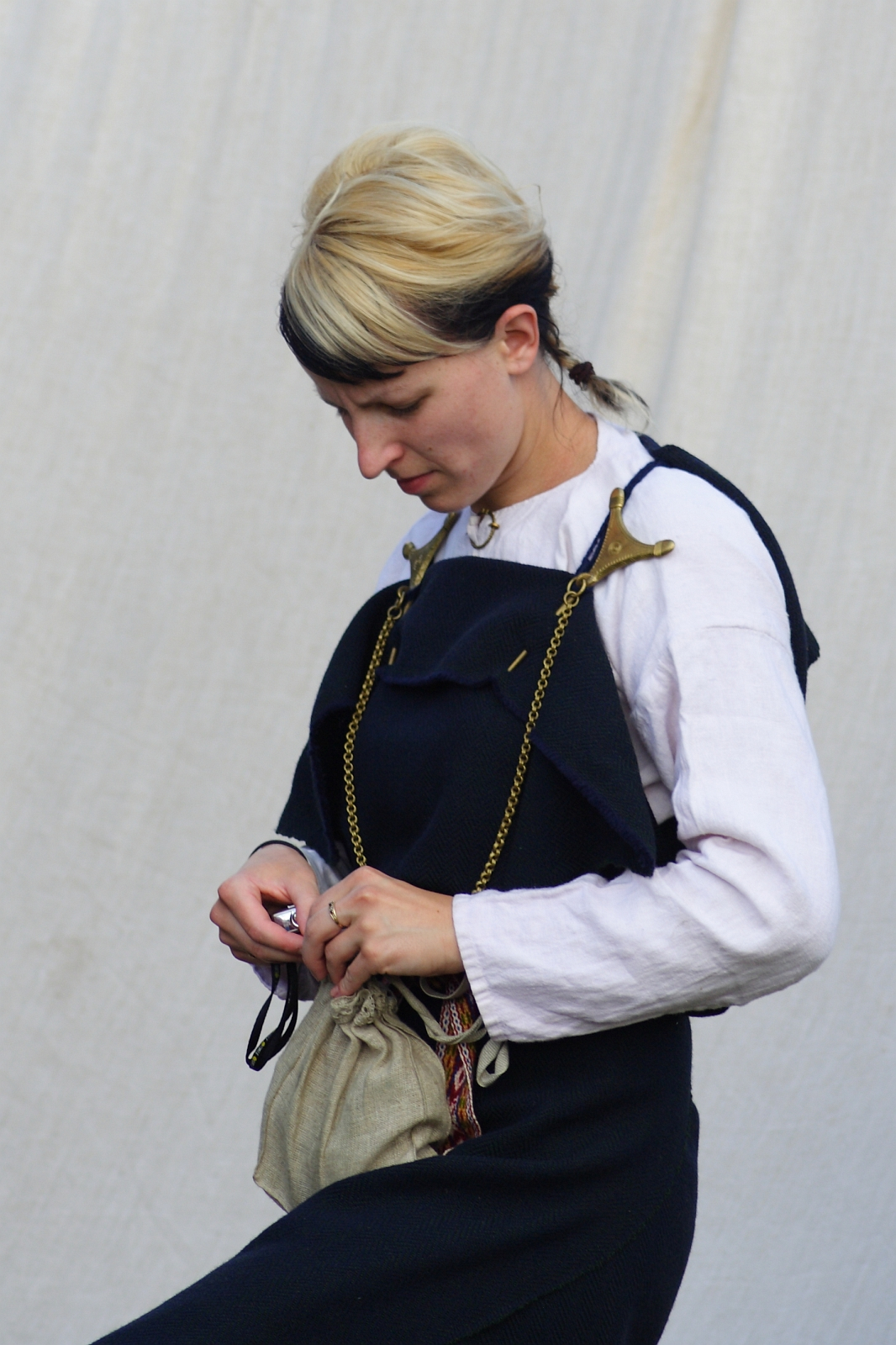 A person with short hair, half blonde, half black, wears a white long-sleeve shirt under a dark blue garment fastened with two golden brooches. They stand against a light-colored background, looking down while opening a small beige pouch hanging around their neck—reminiscent of the Semigallian days.