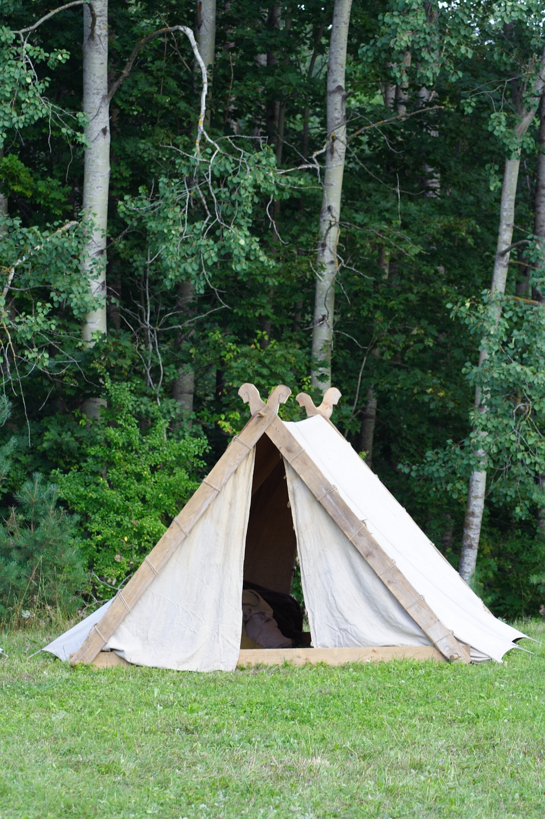 A small, white canvas tent is set up on a grassy area, with dense green trees in the background. The tent has a wooden frame with decorative carvings at the top, and its entrance is open. The simple, rustic setup suggests an outdoor event or perhaps Semigallian Days 2009 in a forested area.