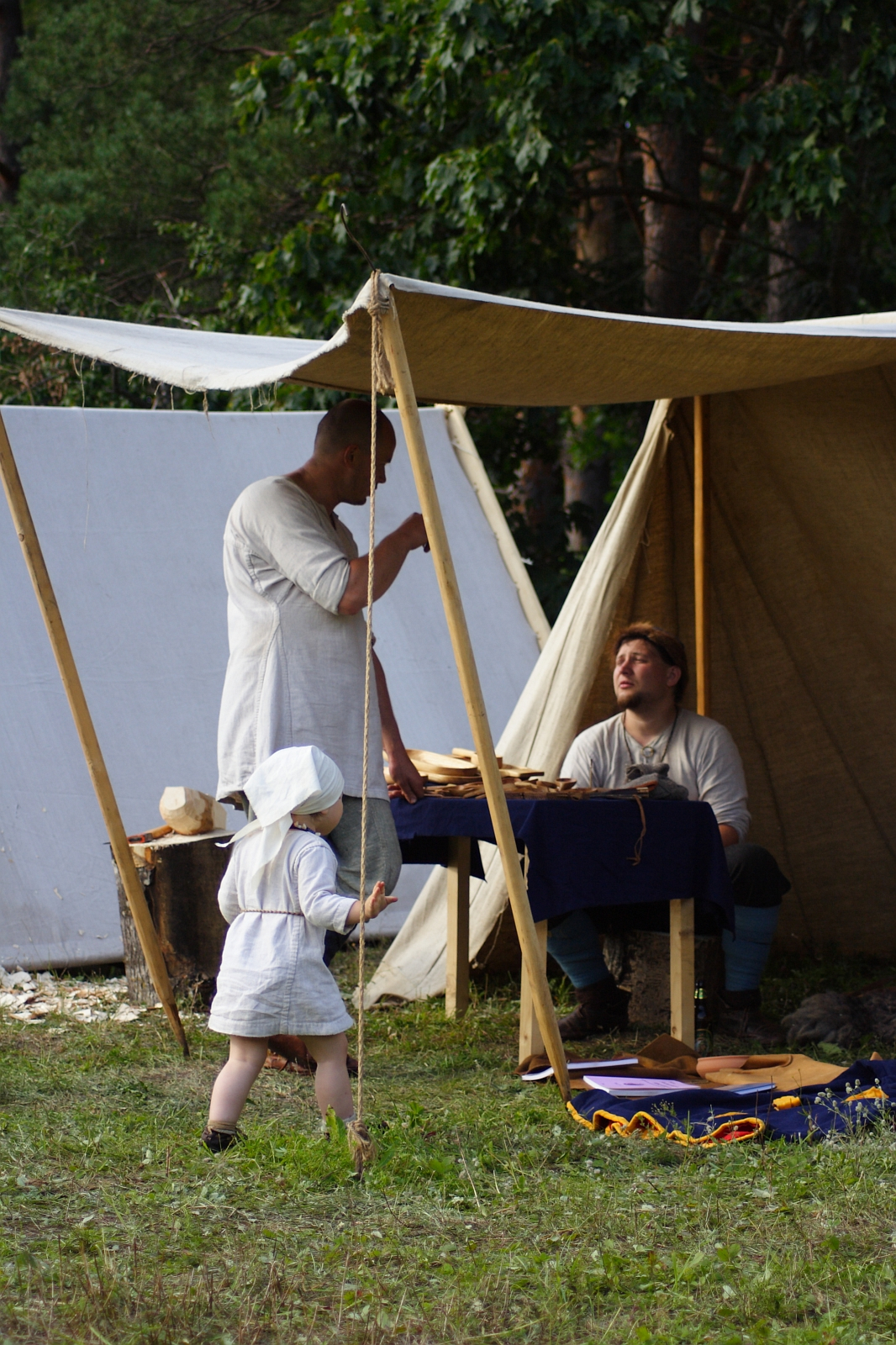 A historical reenactment scene from the 2009 Semigallian Days shows two adults and a child in period clothing under a canvas tent. One adult stands, facing another seated at a table with various wooden tools. The child, wearing a white bonnet and dress, appears to approach the table. The backdrop is a forested area.