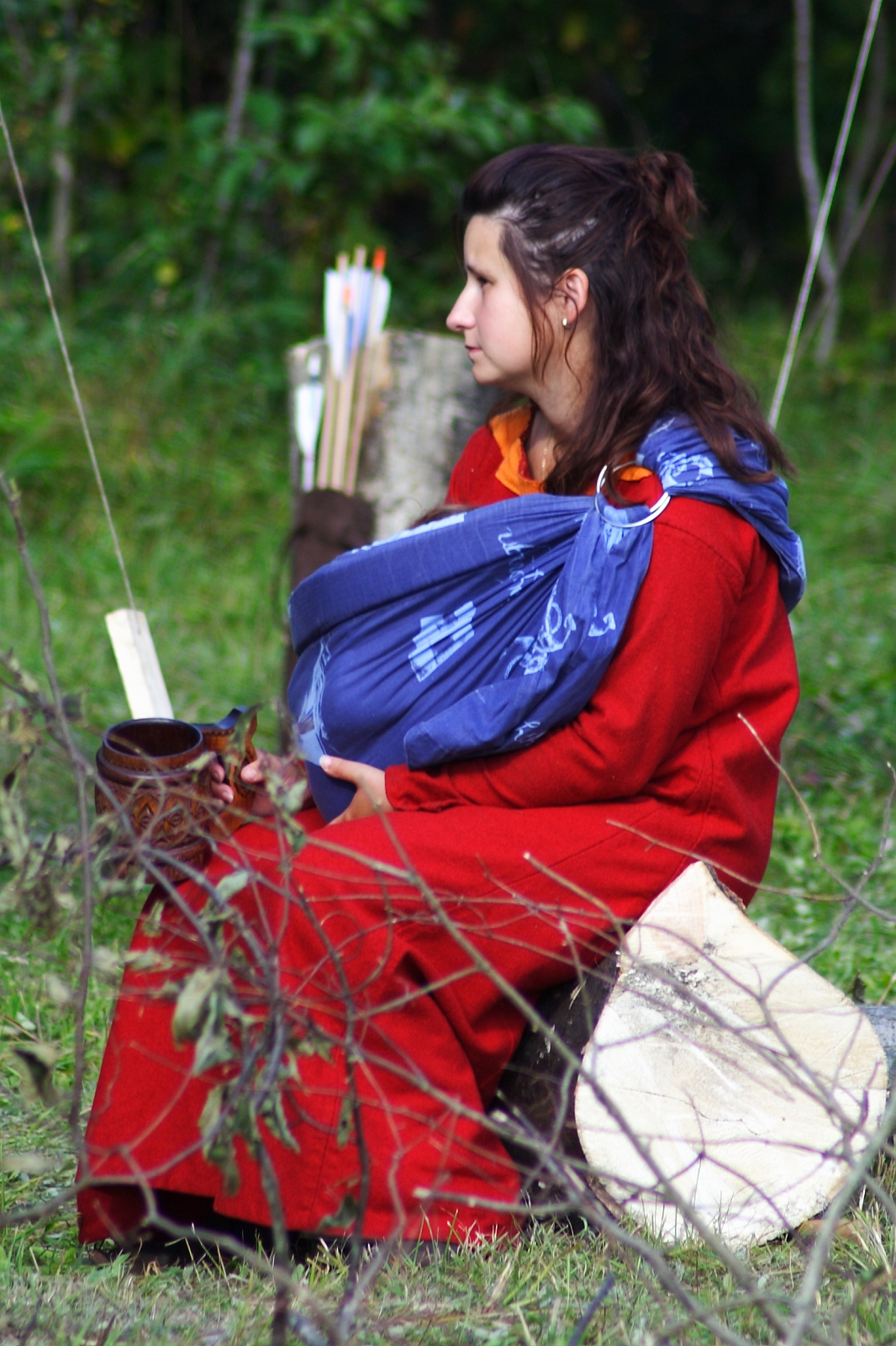 At the 2009 Semigallian Days, a woman in a red dress sits on a log outdoors, holding a mug in one hand and a baby wrapped in a blue cloth carrier in the other. The background features greenery, a wooden stump, and arrows in a quiver. She appears to be gazing into the distance.
