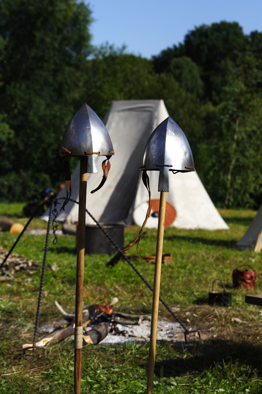 Two medieval helmets on wooden poles stand in the foreground of a historical reenactment campsite. In the background, there are canvas tents and trees, with a small campfire burning near the center. Various camp supplies are scattered around, suggesting an active campsite reminiscent of Semigallian Days 2009.
