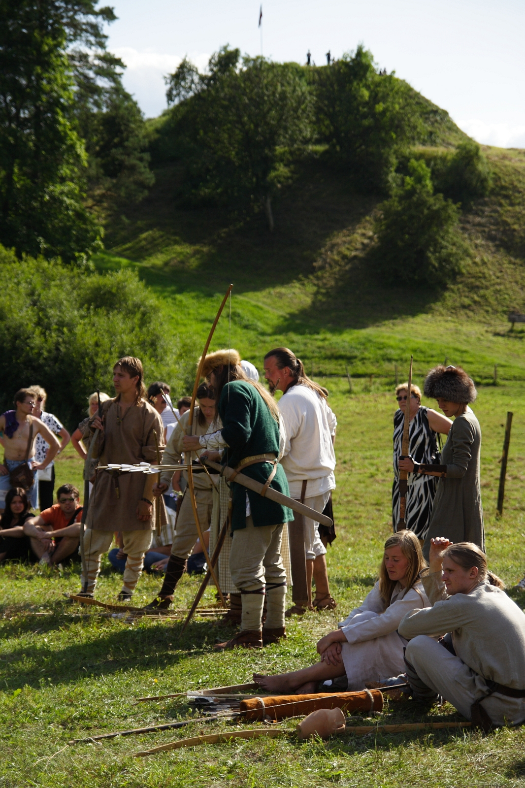 A group of people dressed in medieval clothing gather outdoors on a grassy field by a hillside during the Semigallian Days 2009. Some are standing with bows and arrows, while others are sitting on the grass. Behind them, spectators watch the scene. Trees and a small hill with a flag flying on top are in the background.