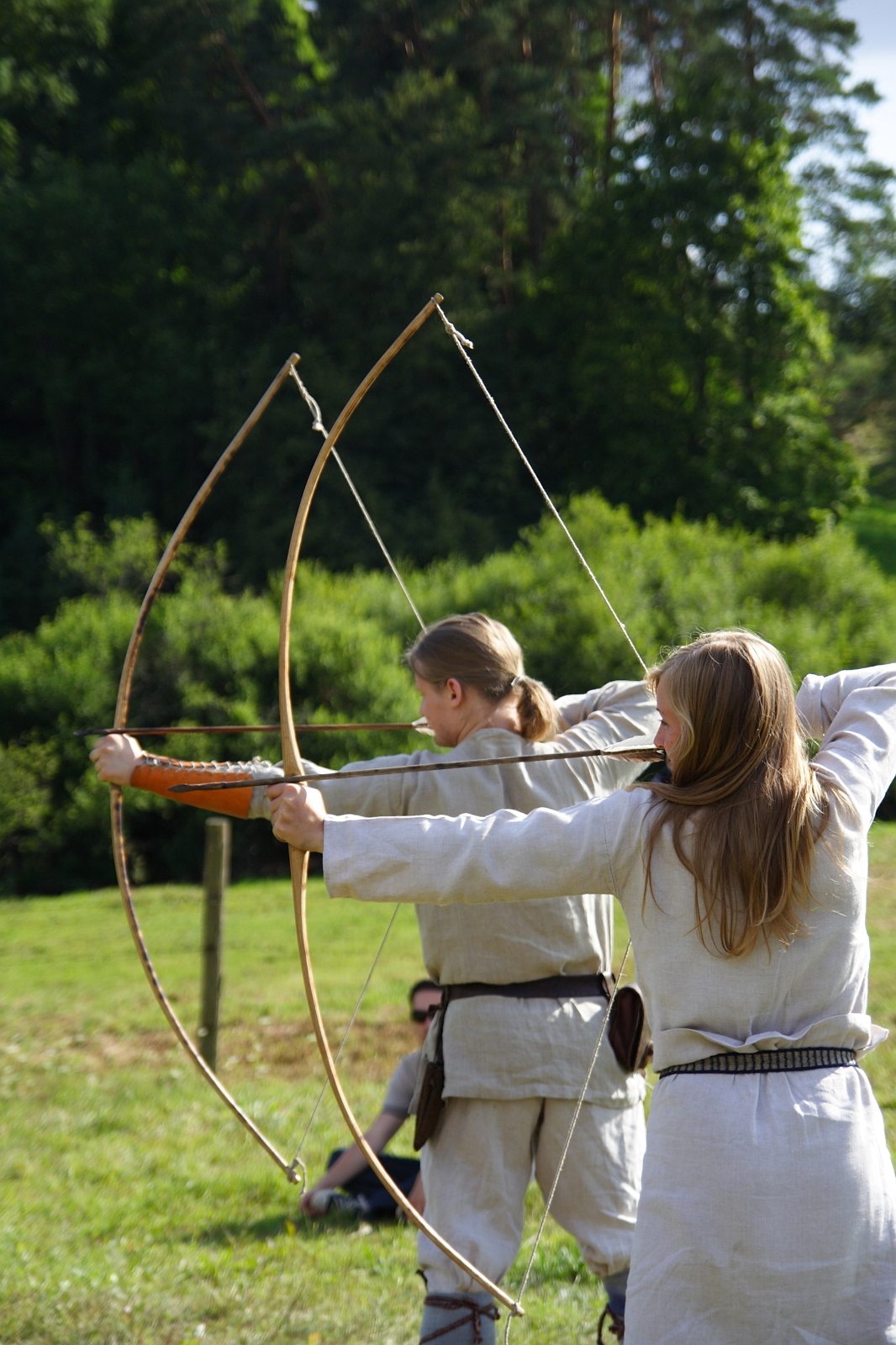Two individuals dressed in Semigallian medieval-style clothing are outdoors, standing on a grassy area. They each hold traditional wooden bows, drawn back in an archery pose, ready to release arrows. A lush, green forest is visible in the background, with sunlight casting bright highlights.