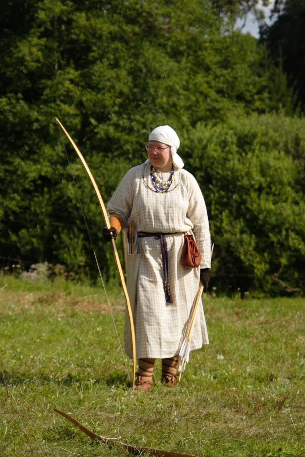 A person dressed in historical Semigallian medieval attire stands in a grassy field with a wooded background. They wear a checked tunic, a headscarf, and hold a longbow. A quiver with arrows is slung over one shoulder, and a brown pouch is attached to their belt. The scene is bright and green.
