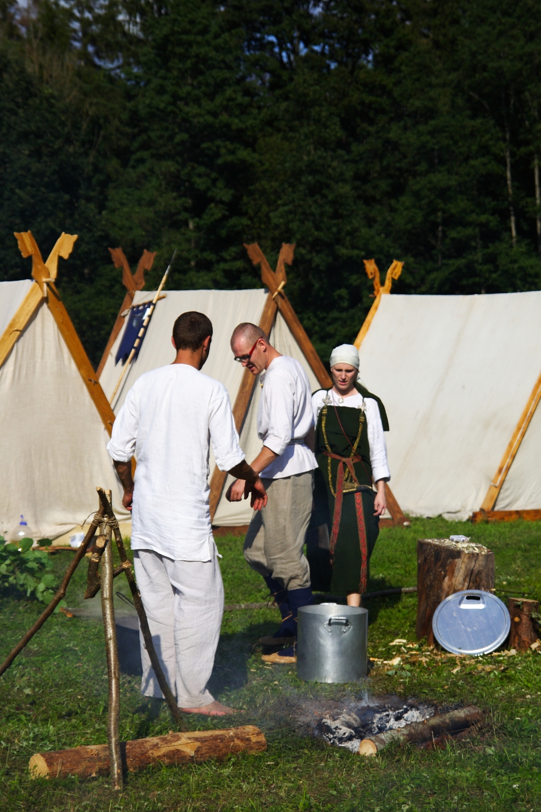 In an outdoor camp with white tents, three people in medieval attire stand by a campfire with a metal pot over it during the 2009 Semigallian Days. One person tends to the fire, while another, dressed in a green tunic and white headscarf, watches. Logs and a shield lie on the grass beside them. Dense trees are in the background.