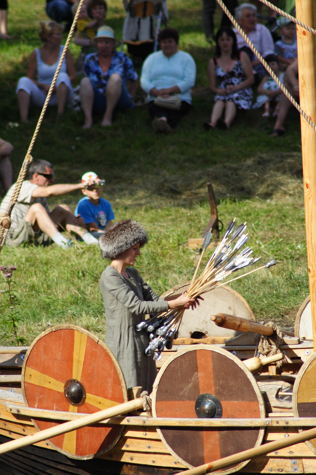 A person dressed in historical attire, wearing a fur hat, is holding a bundle of arrows while standing on a wooden Viking ship during Semigallian Days 2009. The ship is equipped with round shields painted in red and yellow stripes. Spectators, seated on grass in modern clothing, watch the scene from the background.