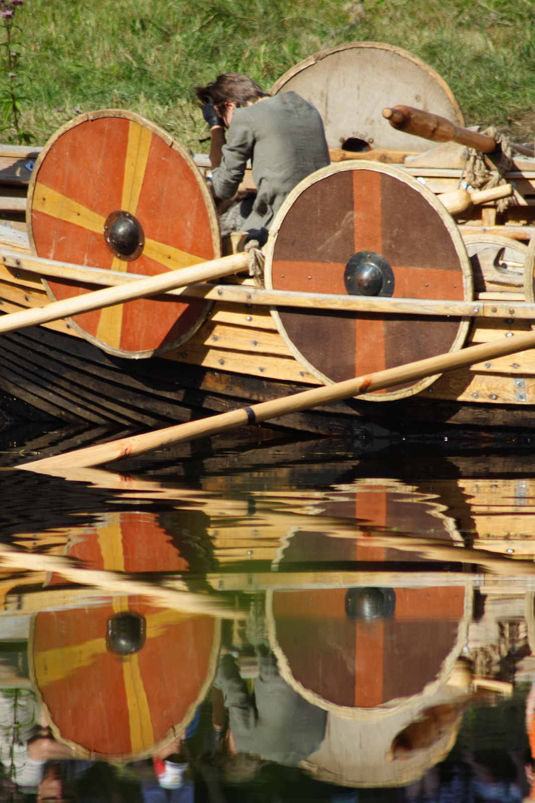 A person sits on the side of a Viking-style wooden boat, adorned with round shields featuring red and yellow patterns. Reflected in the calm water below, the scene captures the essence of the Semigallian festival. The background consists of grass and a wooden pole from the 2009 event.