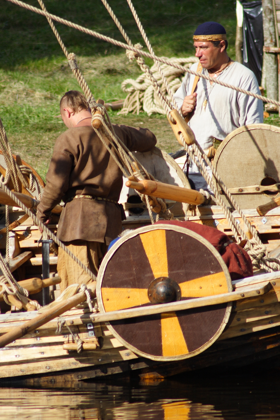At Semigallian Days 2009, two men dressed in historical clothing stand on a wooden boat. One man, facing away, wears a brown tunic while the other, in a white tunic with a black headband, looks forward holding a rope. Visible are circular wooden shields with yellow and brown patterns and various ropes across the boat.