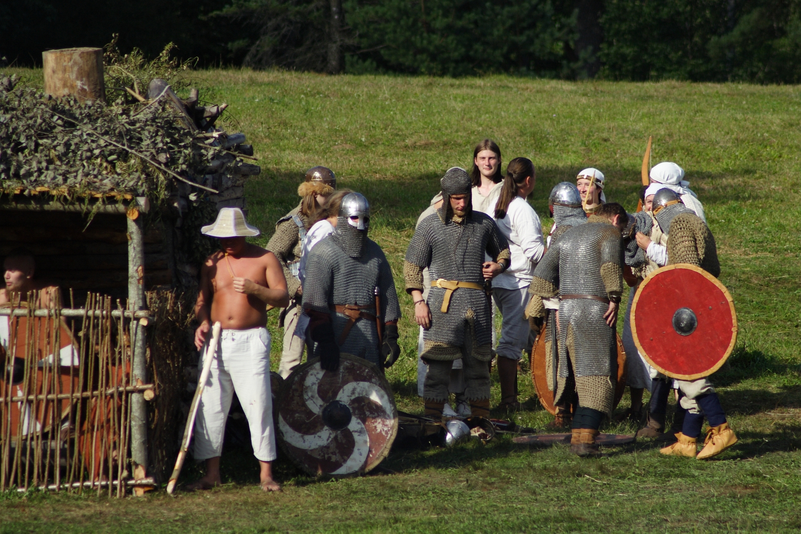 A group of people in medieval costumes gathers on a grassy field at Semigallian Days 2009. Some wear chainmail and helmets with swords and shields, while others are in light tunics. A shirtless man with a white hat stands near a wooden shelter covered with foliage. In the background, trees are visible.