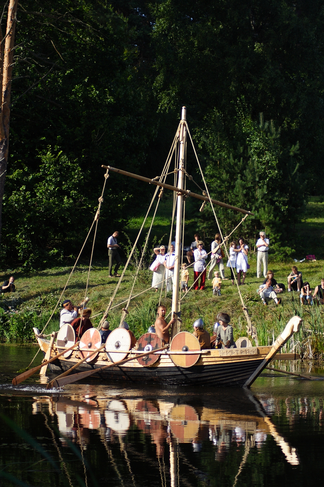 A group of people dressed in historical attire sail a Viking-style wooden boat with round shields on its sides during the Semigallian Days 2009 event. The boat glides on a calm river, reflecting the vessel and passengers. Spectators in varied dress watch from a grassy, tree-lined shore.