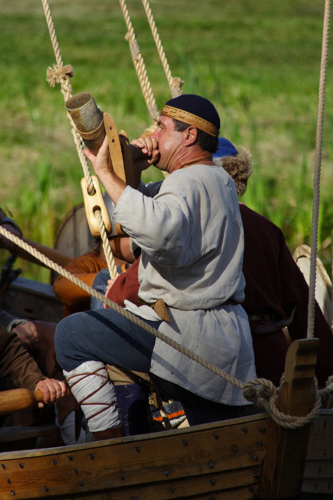 During Semigallian Days 2009, a person in historical attire, wearing a light grey tunic and a navy blue headband, blows into a horn while standing on a wooden boat. Several ropes are visible around the vessel, and other individuals in similar attire are partially seen against the lush greenery.