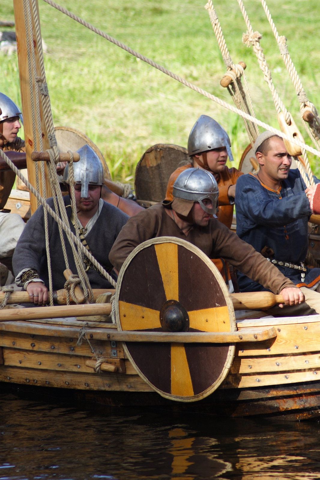 A group of people wearing Viking-style helmets and traditional clothing are on a wooden boat. The boat features oars, ropes, and a shield with a yellow and black design attached to the side. Behind them is a grassy area. The scene suggests a historical reenactment, possibly during Semigallian Days '09.