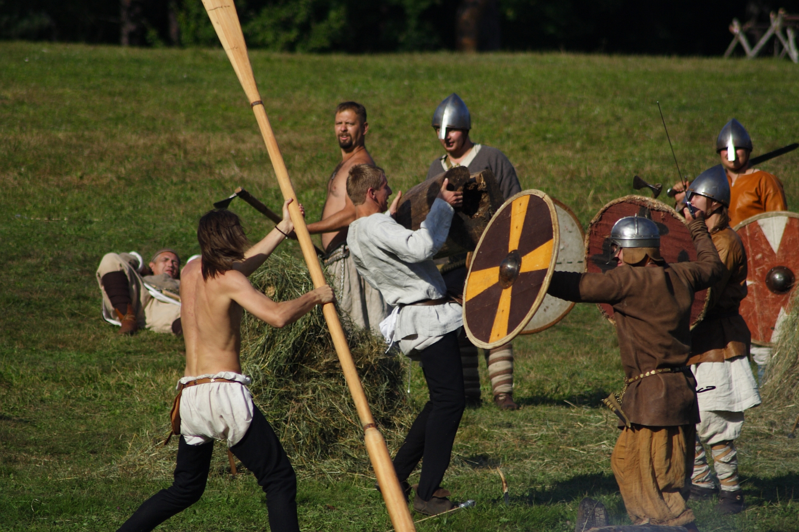A group of people is engaged in a historical reenactment battle on grass during the Semigallian Days. They wear medieval costumes with tunics, helmets, and shields. Two of them clash with swords and shields, while another swings a large wooden paddle. In the background, a few others observe or rest on the grass.