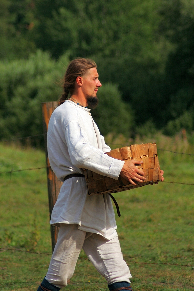 A man with long hair and a beard, dressed in a white linen shirt and pants with a black belt, walks through a grassy area carrying a wooden basket. The background features a wire fence and lush green trees and bushes, evoking the charm of the 2008 Semigallian Days. He seems to be in a rural or historical reenactment setting.