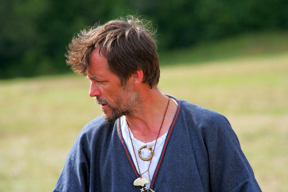 A bearded man with tousled hair, wearing a blue robe over a white shirt and a necklace with charms, stands against a blurred backdrop of greenery and open field. His head is turned to the right, looking downward, conveying a contemplative expression reminiscent of simpler Semigallian days.