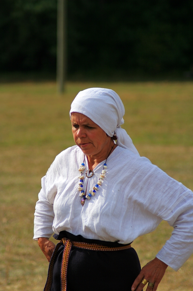 An elderly woman dressed in traditional attire stands outdoors at the 2008 Semigallian Days event. She wears a white headscarf and blouse, with a colorful beaded necklace around her neck. Her hands rest on her hips as she looks down thoughtfully, with a blurred background of grass and trees.