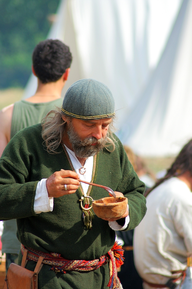 A bearded man in historical attire, including a green tunic and knitted cap, enjoys a meal from a wooden bowl using a spoon during Semigallian days. He stands in an outdoor setting with blurred figures and tents in the background, indicating a possible historical reenactment or festival.