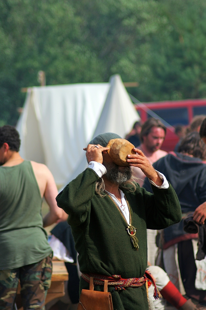 A person dressed in historical attire, including a green tunic and a belt with a pouch hanging from it, drinks from a wooden bowl at the 2008 Semigallian festival. Others in medieval-like costumes are in the background, with tents and greenery also visible, suggesting a historical reenactment or fair.