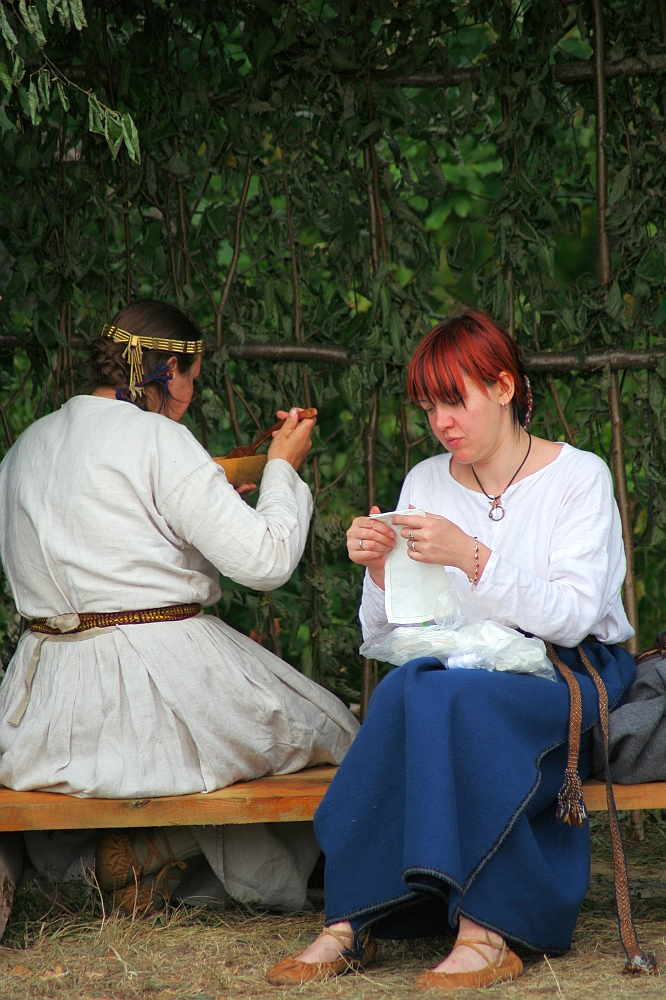 Two individuals in historical clothing sit outdoors on a wooden bench during the 2008 Semigallian Days festival. The person on the left, with braids and a headband, is eating from a bowl. The person on the right has red hair and is focused on sewing white fabric. They are under a leafy canopy, with a bag resting beside them.