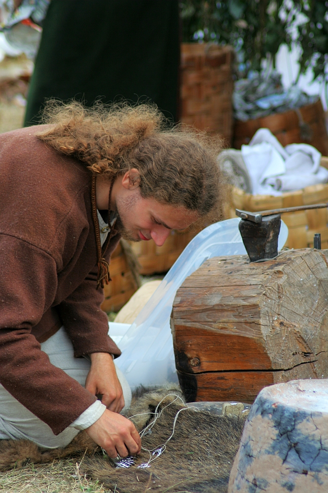 A person with long, curly hair, wearing a brown garment, is sitting on the ground and working with tools and materials, possibly engaging in a craft or historical reenactment. They are surrounded by wooden items, fabric, and other crafting materials at what appears to be the Semigallian festival.
