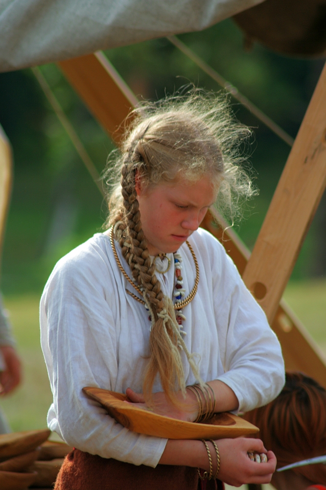 A young person with long, braided hair wears historical clothing and jewelry, including beaded necklaces and bracelets, while holding a wooden bowl. They appear to be participating in Semigallian Days '08, an outdoor historical reenactment or event, with a green, blurry background and wooden structures around them.