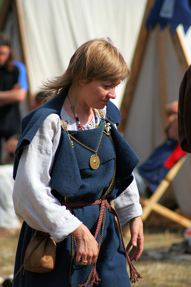 A person with short blond hair is dressed in medieval clothing, featuring a layered blue tunic and white undershirt, accessorized with a leather belt and pouch, necklaces, and a circular pendant. They are outdoors at Semigallian Days '08, with blurred tents and people in the background from the historical reenactment event.