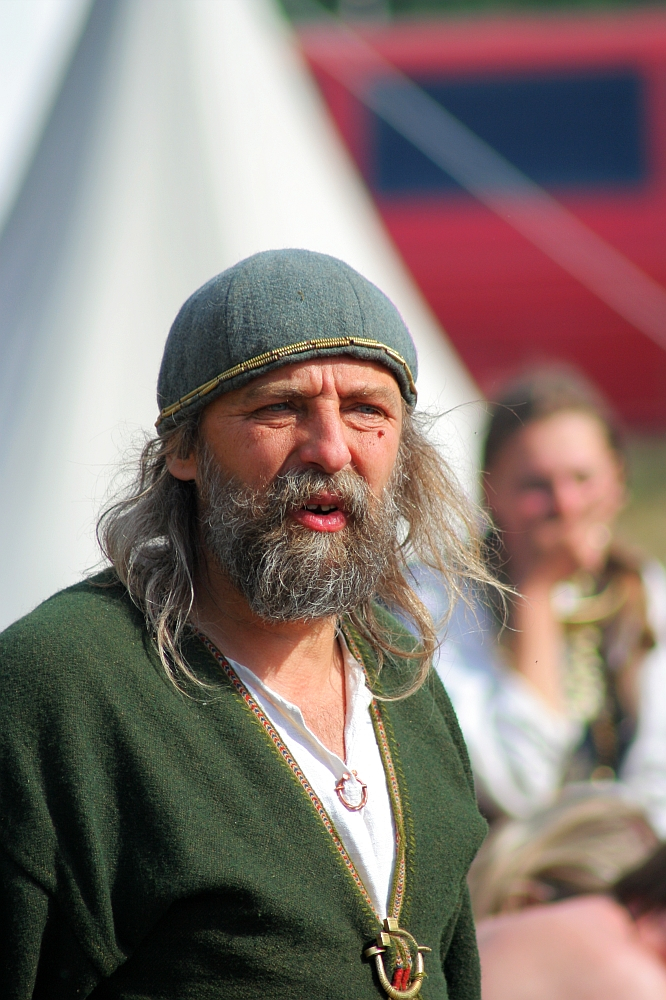 A man with long gray hair and a full beard, wearing a gray cap and green tunic with a white undershirt. He has a necklace with a metal pendant. The background shows a blurred figure of another person and a conical tent with a red structure partially visible at the Semigallian Festival, part of the 2008 events.