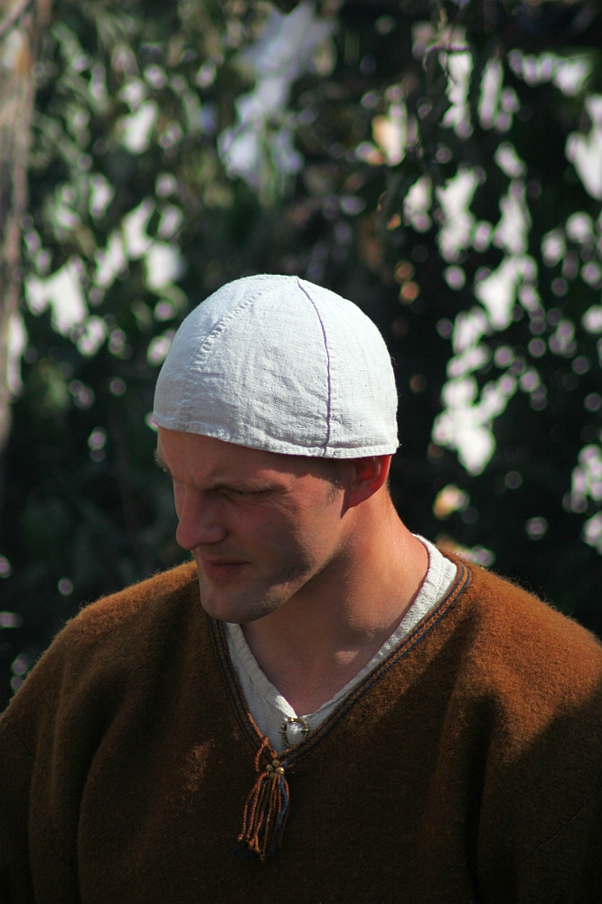 A person wearing a white cap and a brown tunic with a tassel stands outdoors at the 2008 Semigallian Days festival. The individual has a focused expression, and the background is filled with blurred greenery. The tunic appears to be made of coarse fabric, typical of historical reenactment clothing.