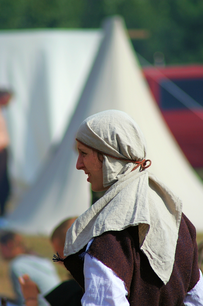 A person is dressed in medieval-style clothing, featuring a light headscarf and a dark tunic with a white undershirt. The background shows a blurred outdoor setting with a conical tent and people who appear to be part of the 2008 Semigallian Days historical reenactment or festival.