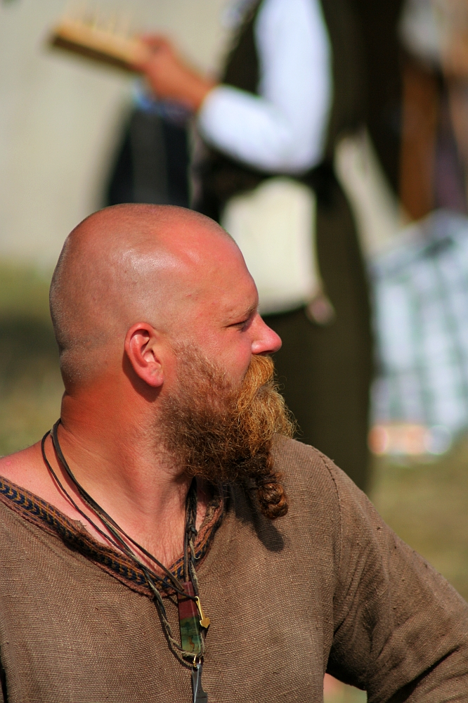 A bald man with a thick red beard is dressed in a brown tunic and adorned with necklaces, including one with a large pendant. He looks to his left, likely reminiscing about Semigallian days. The background is out of focus, showing another person holding an object as sunlight illuminates his face and shoulders.