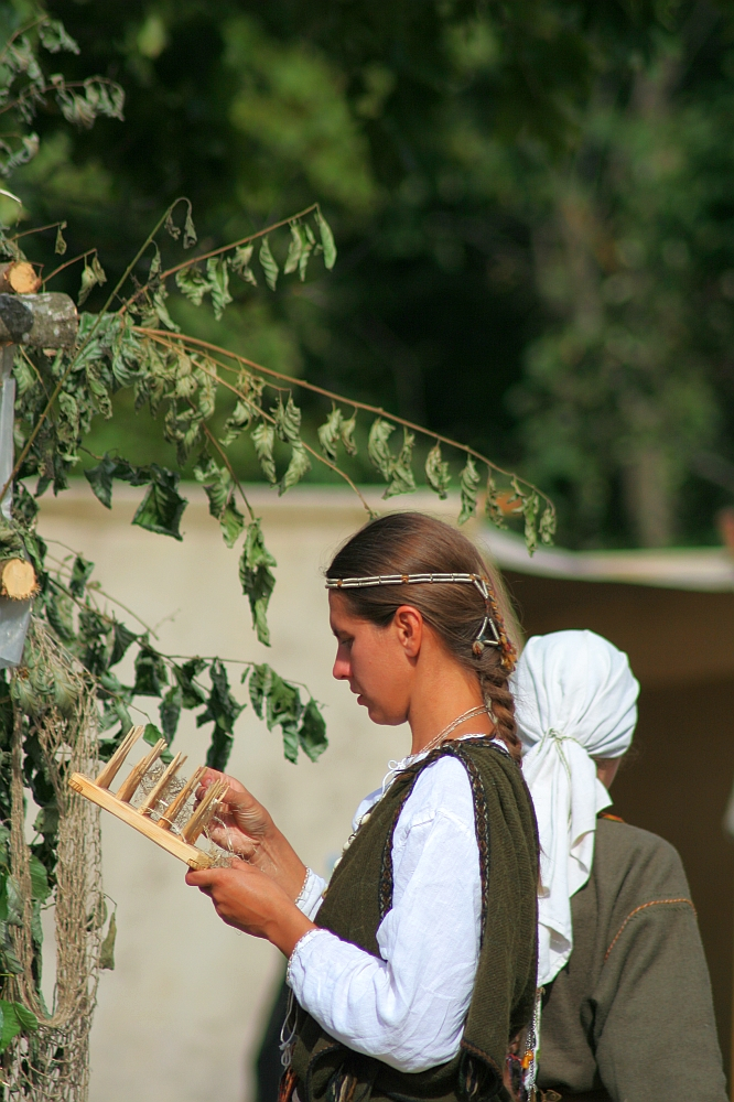 A woman dressed in traditional Semigallian attire, with a white blouse and dark vest, stands outdoors focused on weaving or crafting with a wooden tool during the 2008 Semigallian Days. Her hair is tied back in a ponytail. Another person, partially visible, is standing nearby wearing a white headscarf. Trees and greenery are in the background.