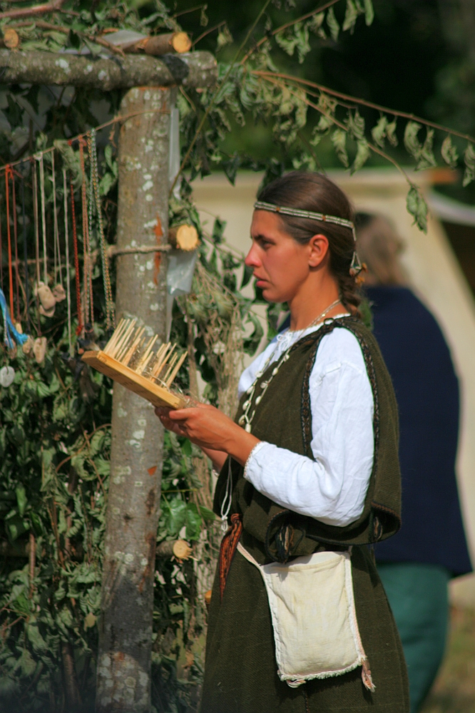 A woman dressed in medieval-style clothing is seen weaving or inspecting threads on a wooden loom at the Semigallian Days festival. She has long hair tied back with a headband, wearing a white undergarment and dark overdress, with a pouch at her waist. Behind her are hanging necklaces and greenery, part of the 2008 historical reenactment setup.