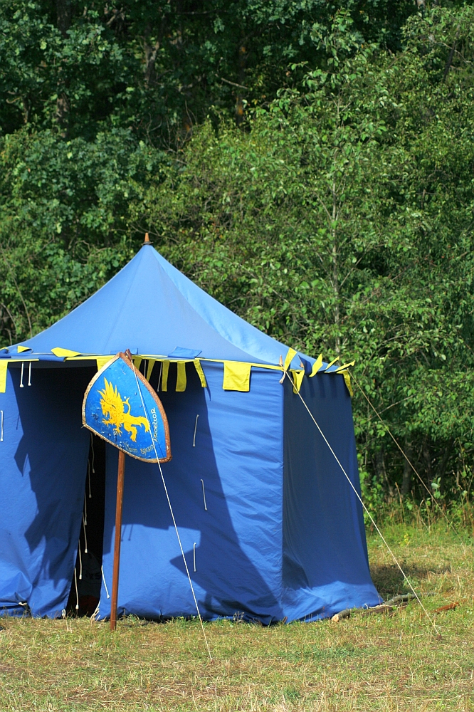 A blue medieval-style tent with yellow trim stands on grassy ground. The tent has an opening in the front, revealing a dark interior. Beside the entrance is a blue shield featuring a yellow griffin, reminiscent of the days gone by. The background consists of dense green foliage.