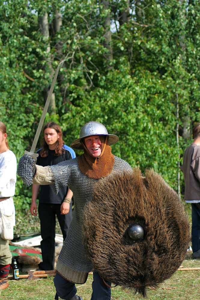 A person dressed in medieval armor, including a chainmail shirt and a metal helmet from the 2008 Semigallian Days, is smiling and wielding a sword in one hand while holding a fur-covered shield in the other. They stand outdoors with trees in the background and other people dressed in historical clothing nearby.